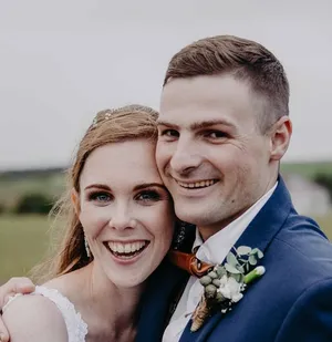 A close-up, heartwarming portrait of a bride and groom on their wedding day.  The bride, with reddish-blonde hair and a white dress, leans against the groom, who wears a navy blue suit and a rustic brown bow tie. They both beam with joy.
