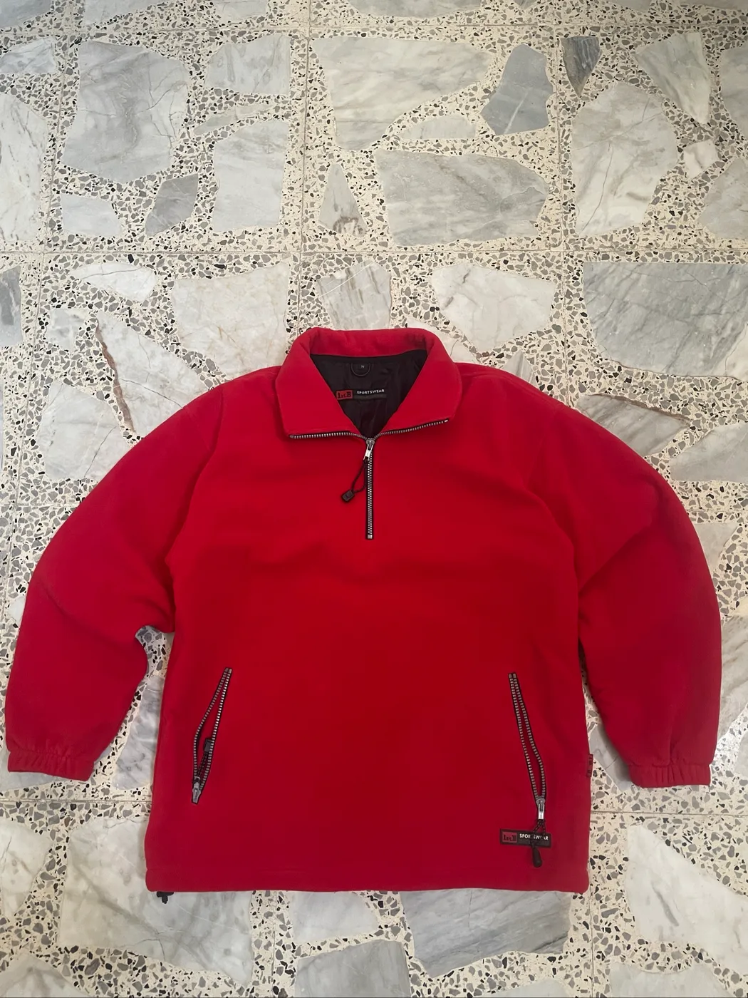 سترة صوفية حمراء (Fleece Pullover) بنصف سحاب وجيوب جانبية بسحاب، وتبدو كقطعة ملابس شتوية كلاسيكية بتصميم مريح.