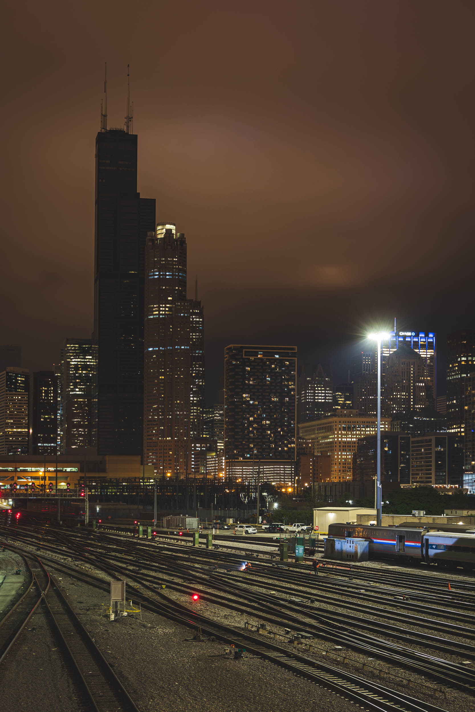 The Sears/Willis Tower without any lights against the Chicago skyline, with two men working on railroad tracks in the foreground below.
