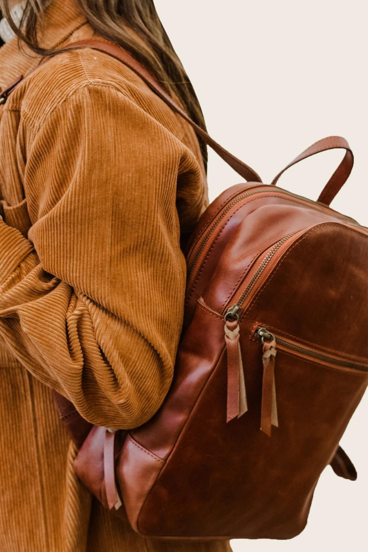 a person models the able liliana everyday leather bag halfway of their shoulder. the bag has two main zipper pockets and one at the front. the whiskey colored pack complements the model's ochre sweater.