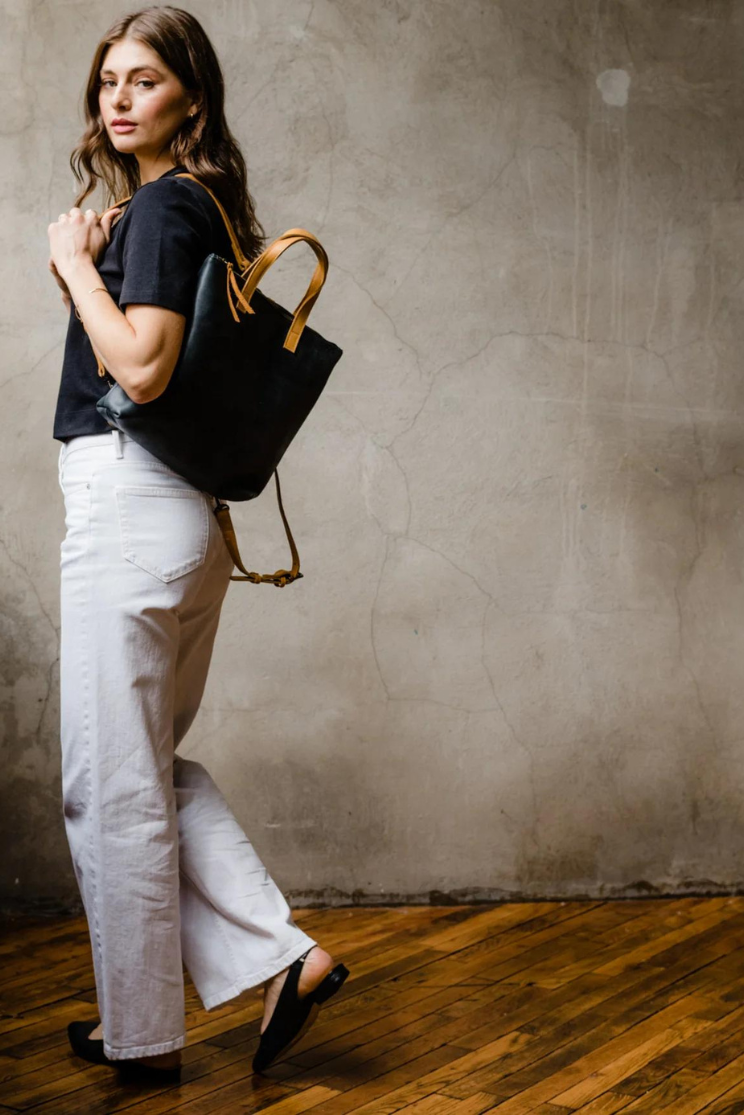 a person models a black abera convertible backpack from a distance half off of her shoulder. it has brown leather straps on the sides and the top. the model wears a black tee with white pants and looks at the camera with her body facing left.
