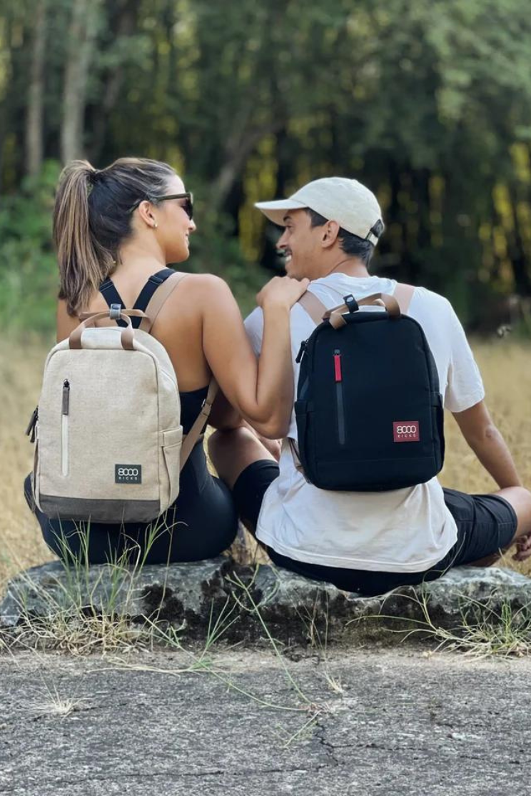 two people model 8000 kicks small backpacks while sitting in the wilderness. the person on the left has sunglasses, a ponytail, and a tank top. they model a beige backpack and looks to the person on her right with a hand on their shoulder. the other person, wearing a white cap, white shirt, and black backpack, looks to the left.