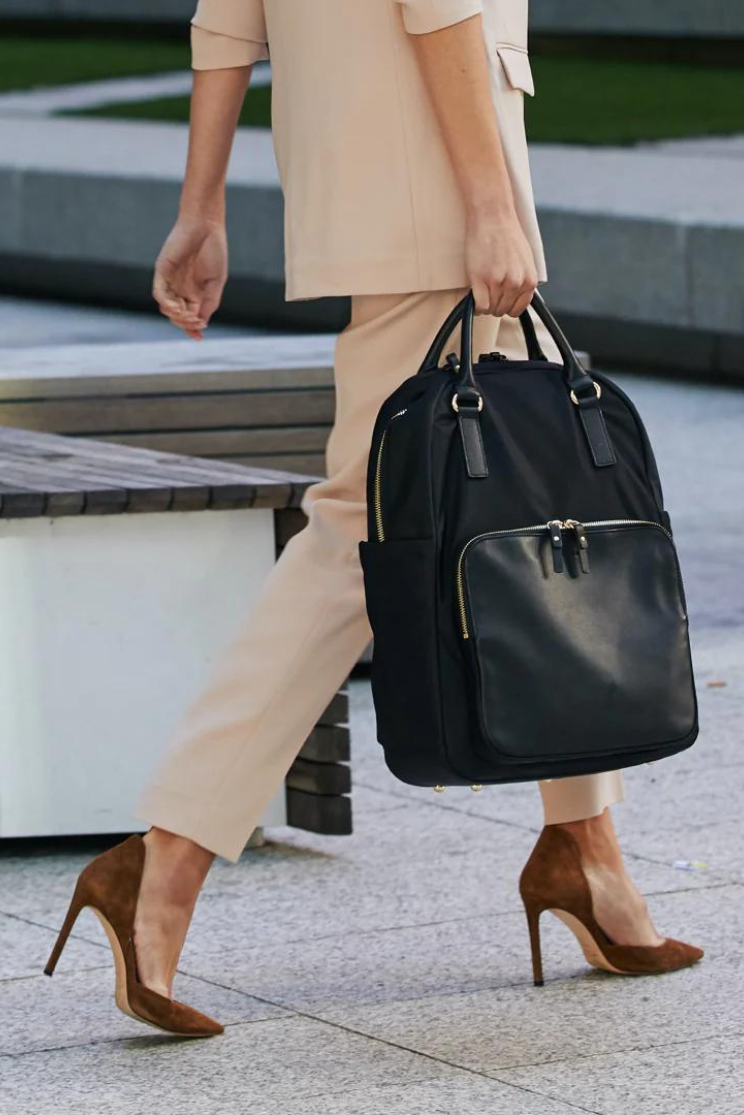 a person in a tan pan suit and brown heels walks with the rowledge backpack by the top straps