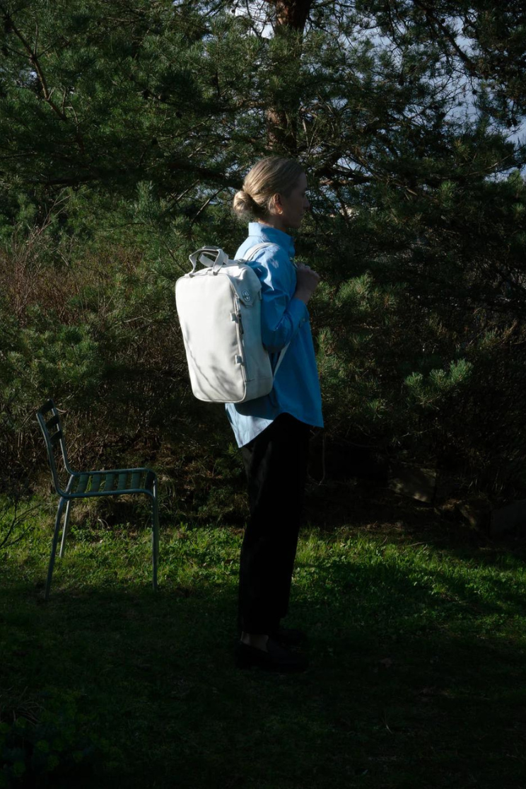 a person models a bananatex daypack in gravel (white.) the bag has no visible zippers from this angle, but has handles on the top. the model faces away from the camera to the right in front of grass and trees.