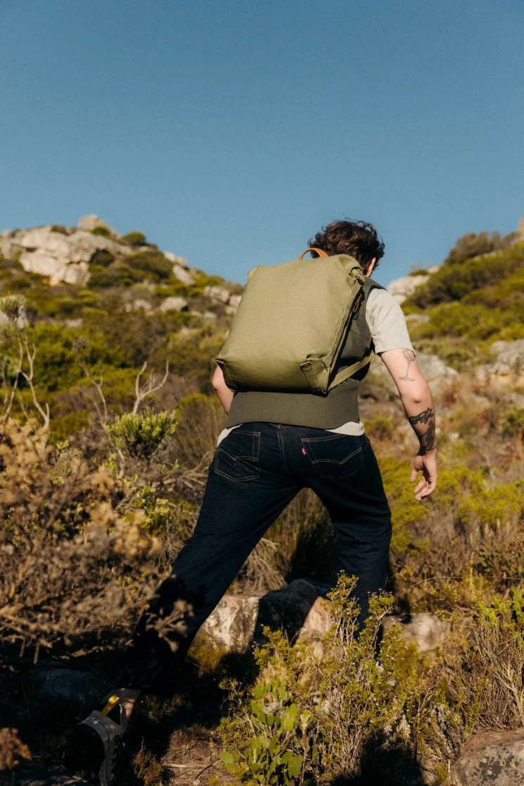 a person models a sage green zip pack while walking up the side of a mountain.
