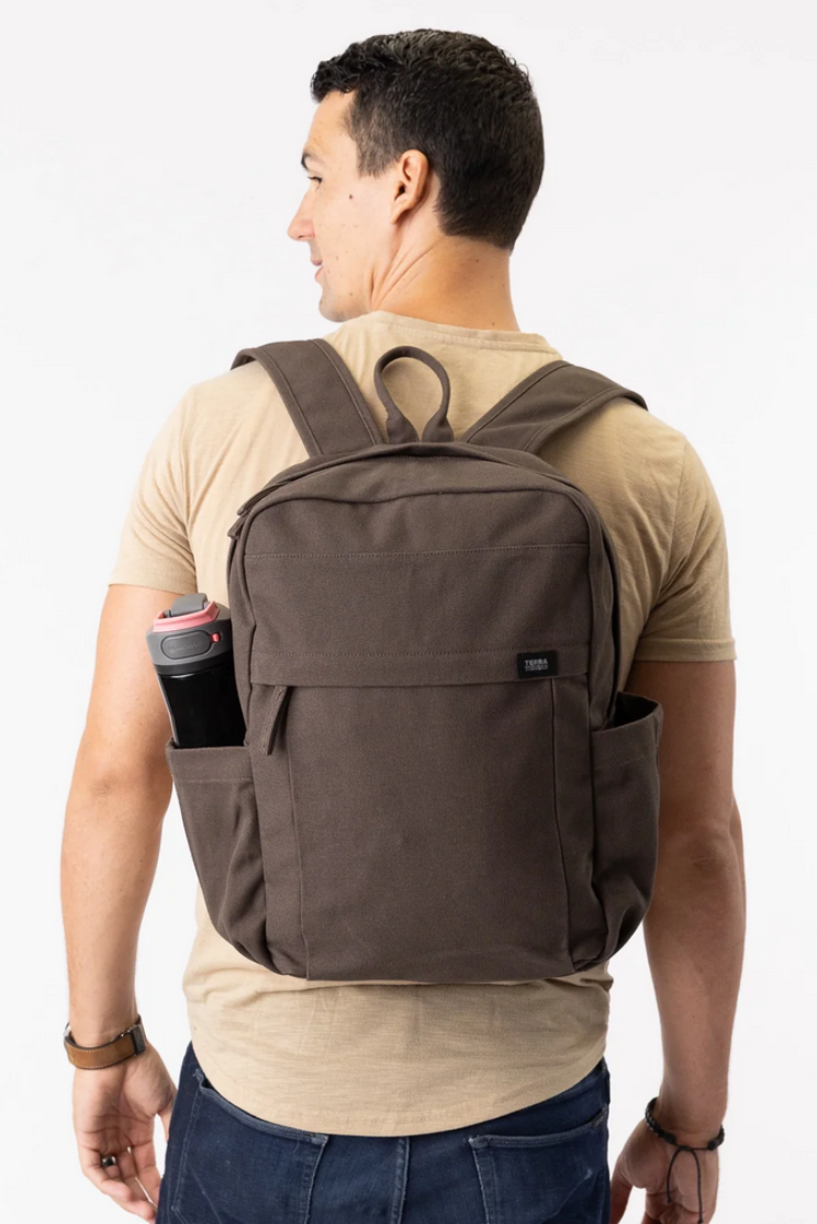 a person models a brown earth backpack from terra thread. it has a water bottle sized pocket on either side and a loop on top