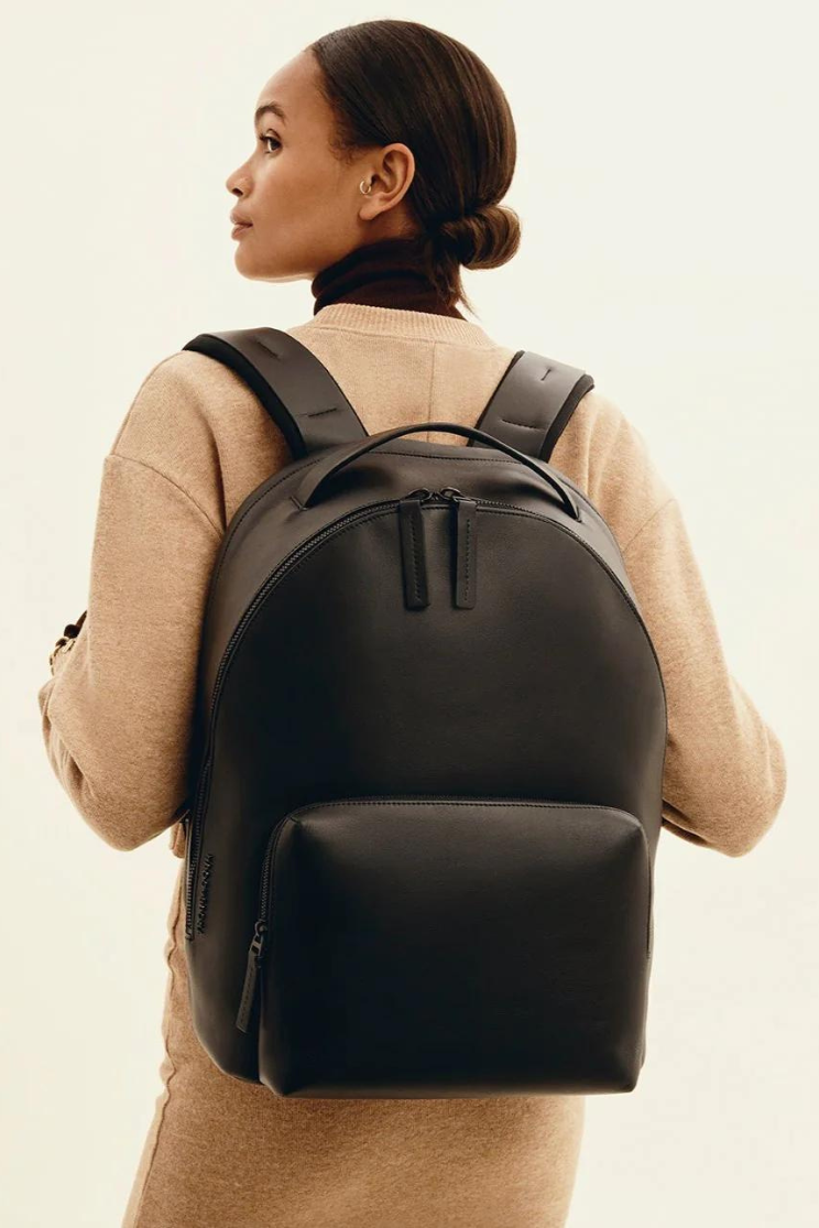 a person models a black generation leather backpack from troubador. it has a large back pocket and two zippers hanging from the top