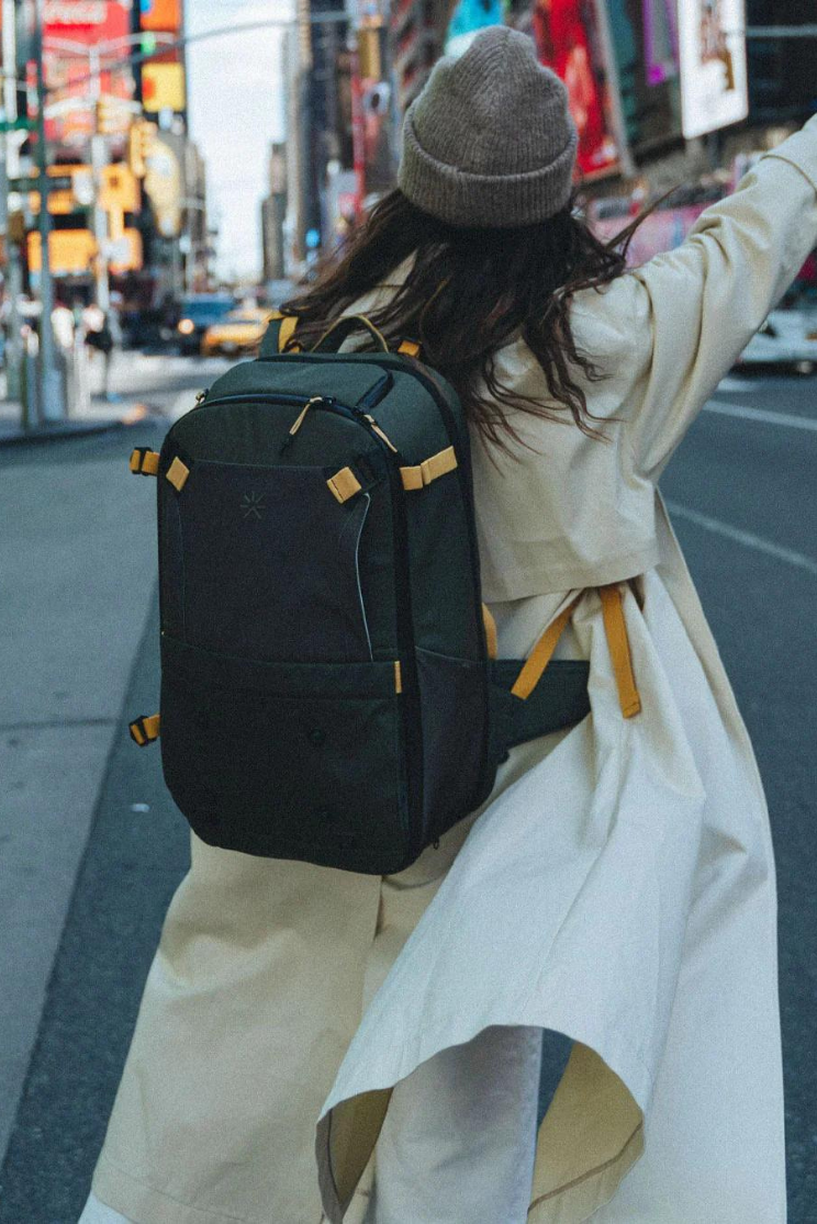a person models a black shell backpack from tropic feel on a city street. they wear a white coat with a gray beanie and wave to the street