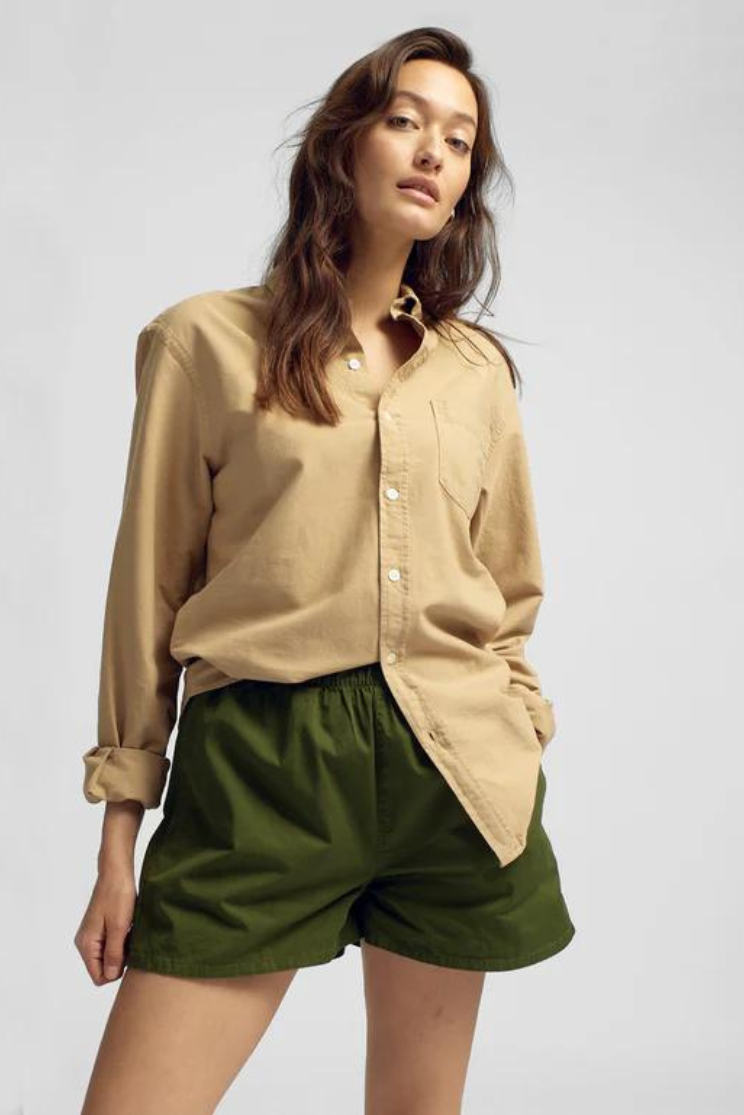 a woman models a tan cotton button down shirt into a pair of green shorts