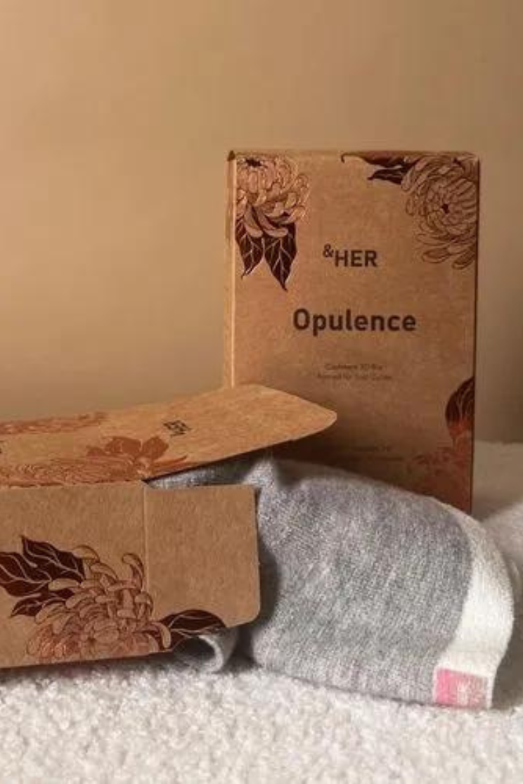 two cardboard packaging boxes, one upright and the other on its side. the boxes are decorated in subtle florals with the words "her" and "opulence"