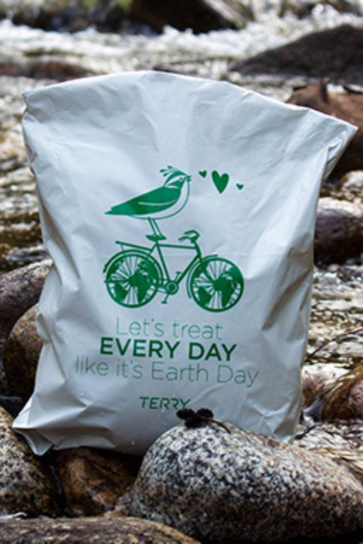 a white recycled poly mailer with a green logo of a bird riding a bike with globes for wheels with the words "let's treat every day like it's earth day" the bag sits on rocks in a creek