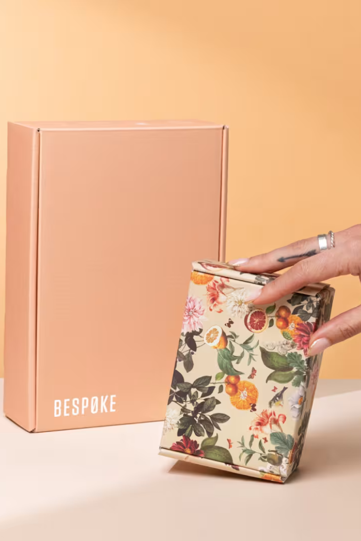 a hand holds a floral cardboard box upright in front of a larger pale pink one that reads "bespoke"