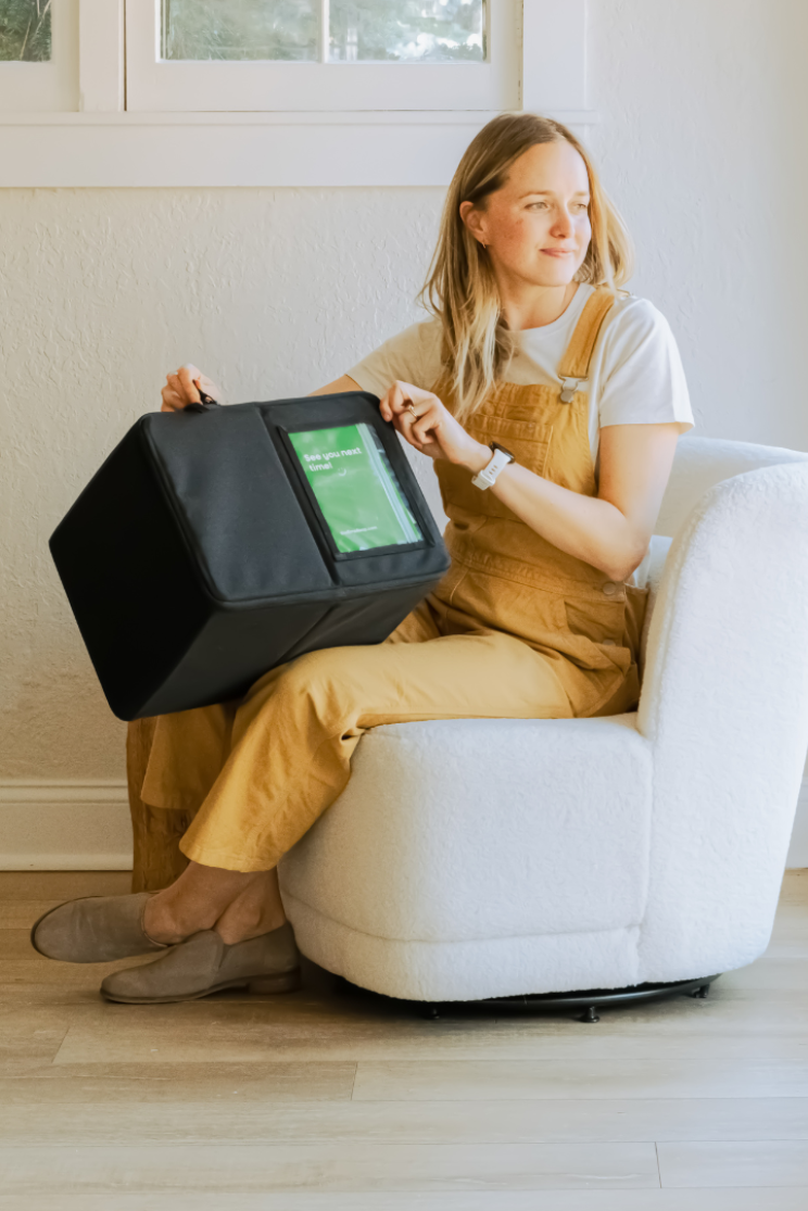 a peron with long blond hair in tan overalls holds a large reusable box mailer while sitting in a chair and looking to the side