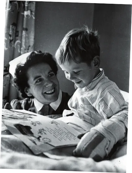 Imagem de uma mulher e uma criança. A mulher usa um chapéu branco sem abas e uniforme com gola abotoada ao redor do pescoço. A mulher olha e sorri para a criança, que está de pijama, sentada em uma cama. A criança tem um livro com mais ilustrações que palavras aberto sobre as próprias pernas e olha sorrindo para esse objeto. A mão esquerda da criança está enfaixada.