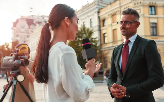Imagem de uma repórter entrevistando um homem. Ao lado deles, um cinegrafista filma a entrevista.