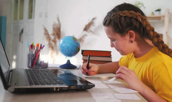 Imagem de uma menina estudando. Ela escreve em um bloco de notas e está sentada diante de um notebook.