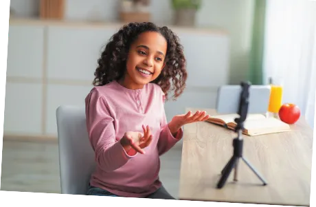 Imagem de uma menina gravando um vídeo com um celular. Ela sorri e gesticula diante do celular, que está preso em um suporte.