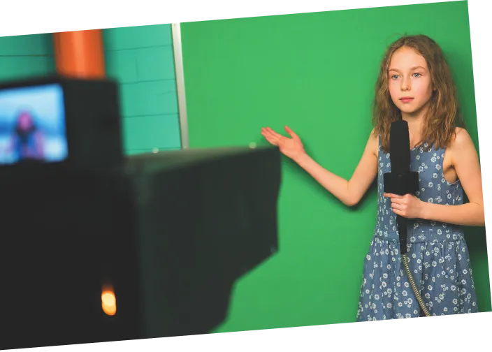 Imagem de uma menina segurando um microfone. Ela está diante de uma câmera de filmagem.