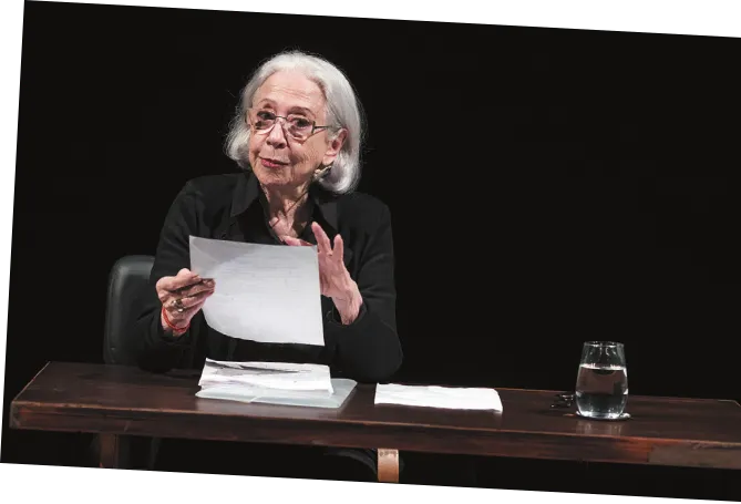 Imagem de Fernanda Montenegro. Ela está sentada diante de uma mesa e lê um texto escrito em folhas de papel.