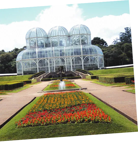 Imagem de uma construção transparente, com estrutura tubular, diversos arcos e três cúpulas. Na frente da construção há um jardim e um chafariz.