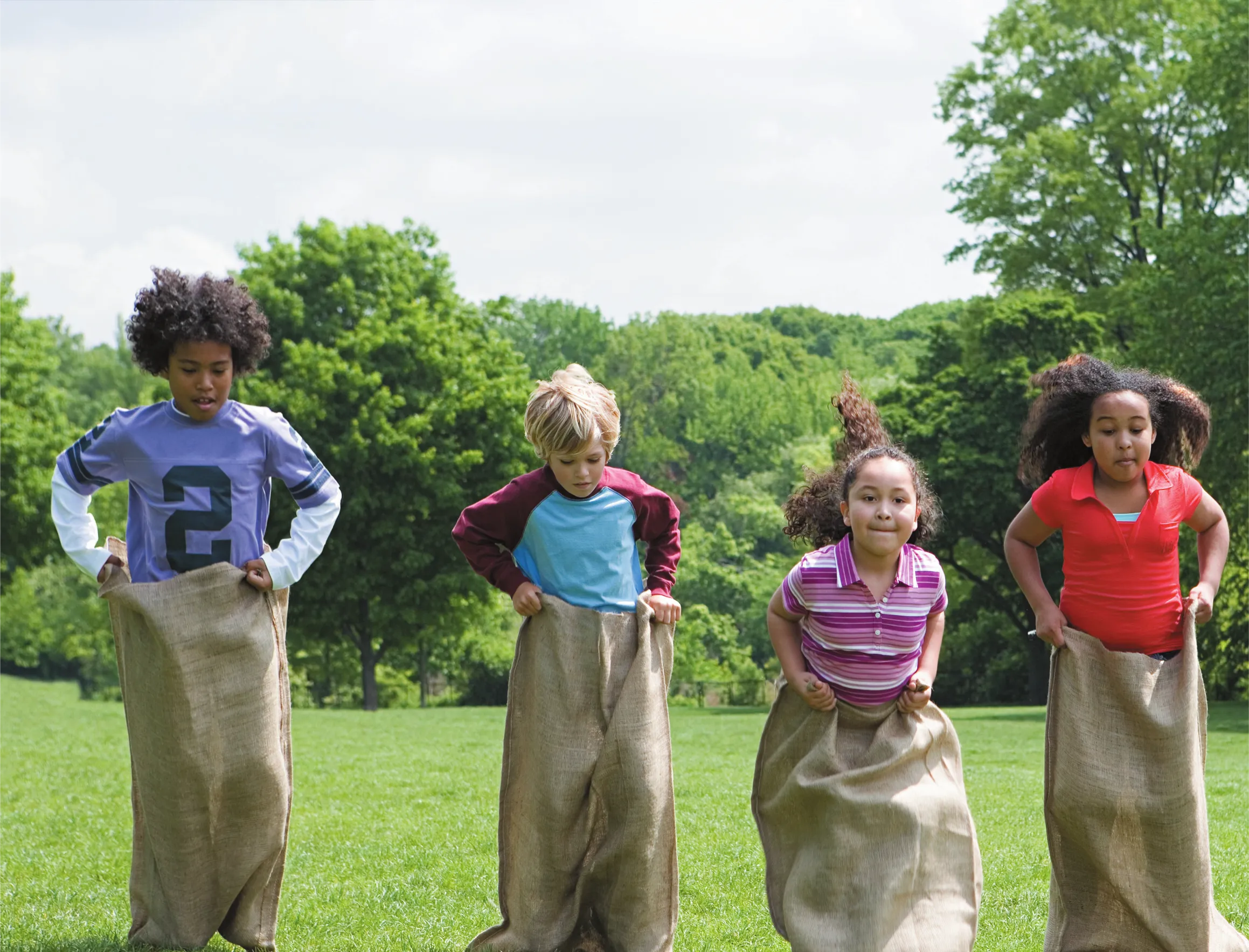 Imagem de quatro crianças brincando. Elas estão em uma área plana gramada e se deslocam pulando. Cada criança está com as pernas dentro de um saco que se estende até a cintura.