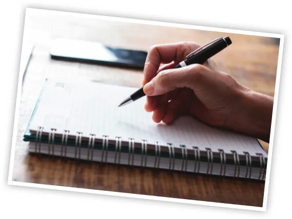 Imagem da mão de uma pessoa segurando uma caneta sobre um caderno aberto.