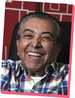 Imagem de Maurício de Sousa. Ele é idoso, usa uma camisa xadrez e está sorrindo.