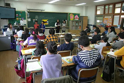 Imagem de sala de aula com alunos sentados em duplas. À frente da lousa, há dois pares de alunos. A professora aparece sentada à esquerda.