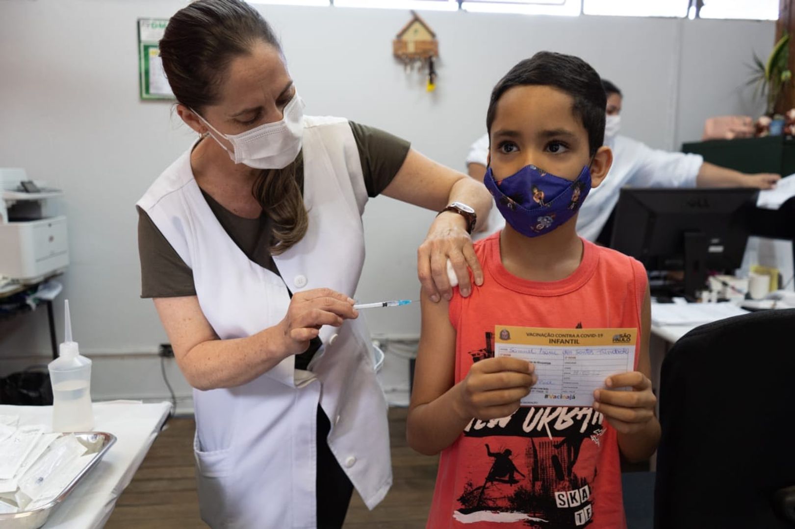 Enfermeira usa máscara, jaleco branco e está vacinando uma criança. A criança é um menino de pele negra, usa máscara de proteção facial, uma camiseta vermelha e está segurando a carteira de vacinação infantil contra COVID-19.