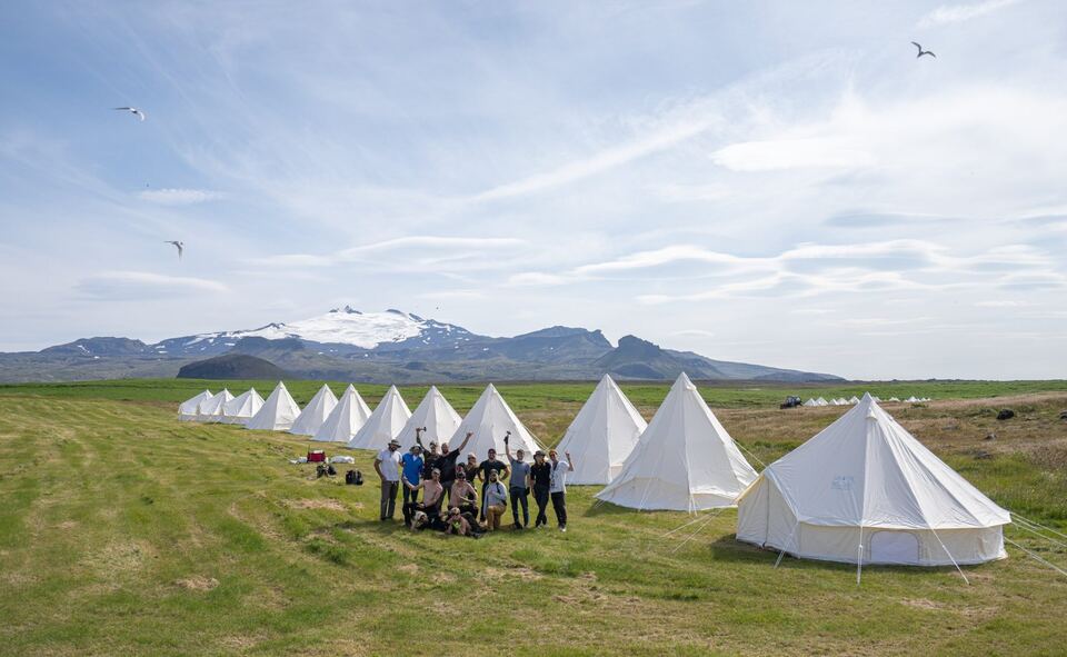 The build crew cheering among rows of white bell tents, with the glacier behind