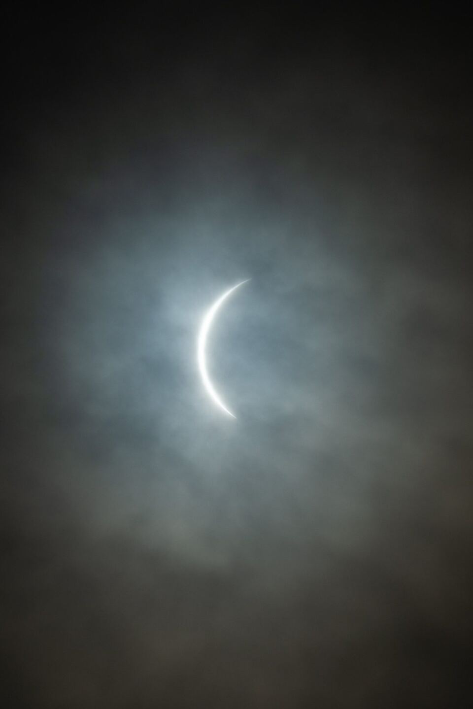 A thin crescent of the eclipsed Sun shining through a break in grey cloud