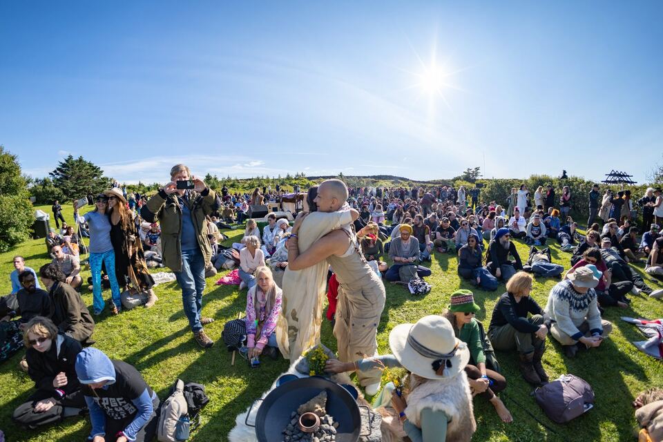 Hundreds of people sitting on the grass in the sun at the opening ceremony, two women embracing