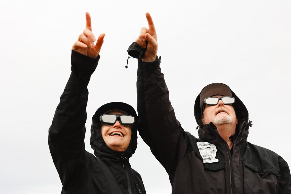 Two Eclipse Seekers in rain jackets and eclipse glasses pointing up at the sky, one laughing