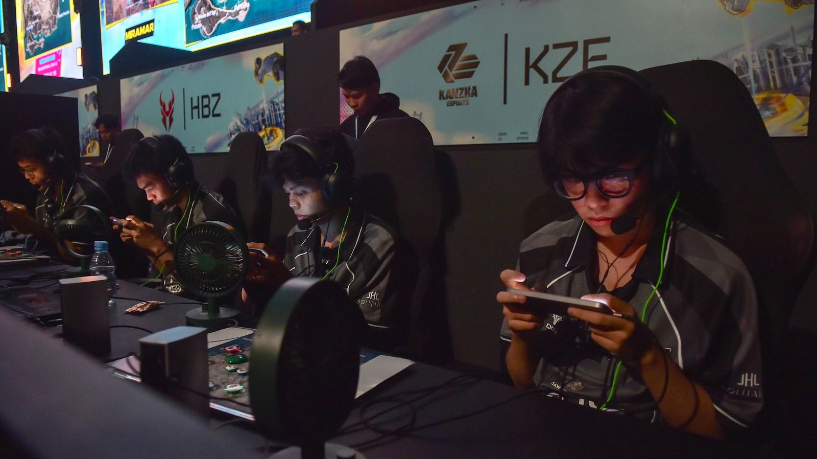 A group of gamers in matching jerseys focus on their mobile devices during an esports event, with screens and fans in the background.