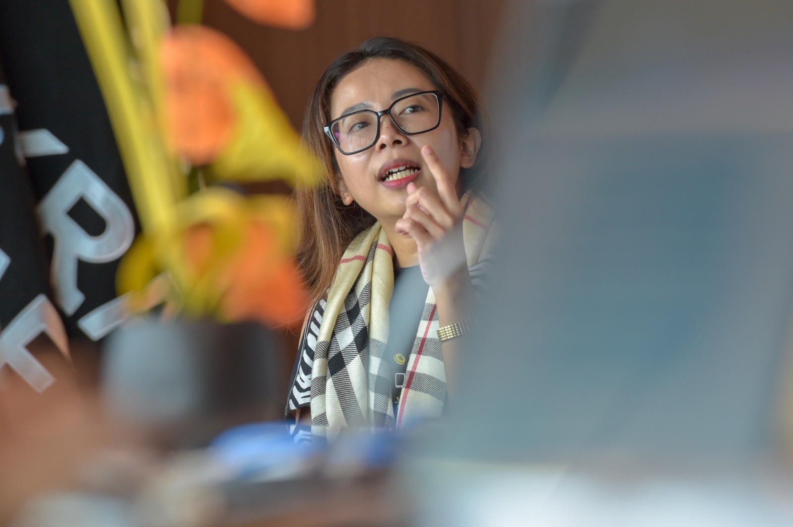 A woman with glasses gestures while speaking, wearing a patterned scarf, with plants and a blurred background.