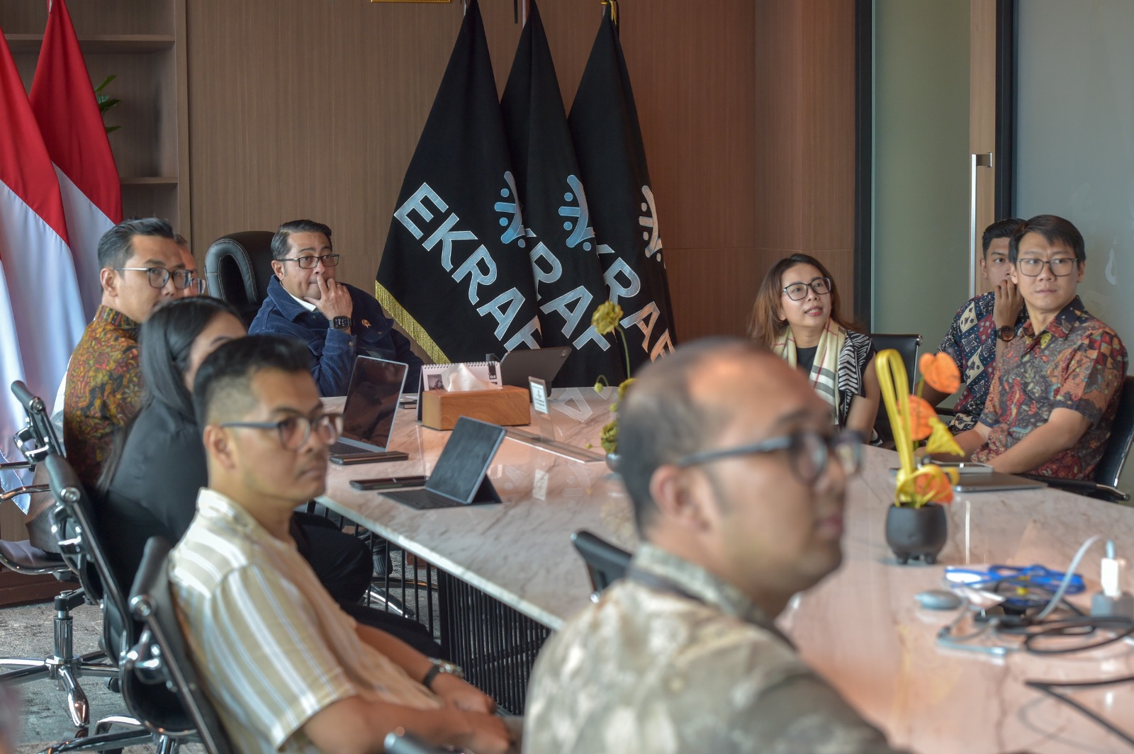 A group of people in a meeting room, seated around a table with laptops, flags in the background, and a floral arrangement.
