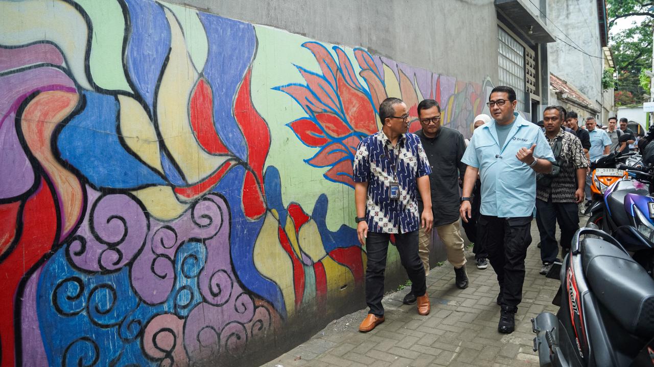 A group of men walks along a colorful mural on a wall, discussing as they pass parked motorcycles in a narrow alley.