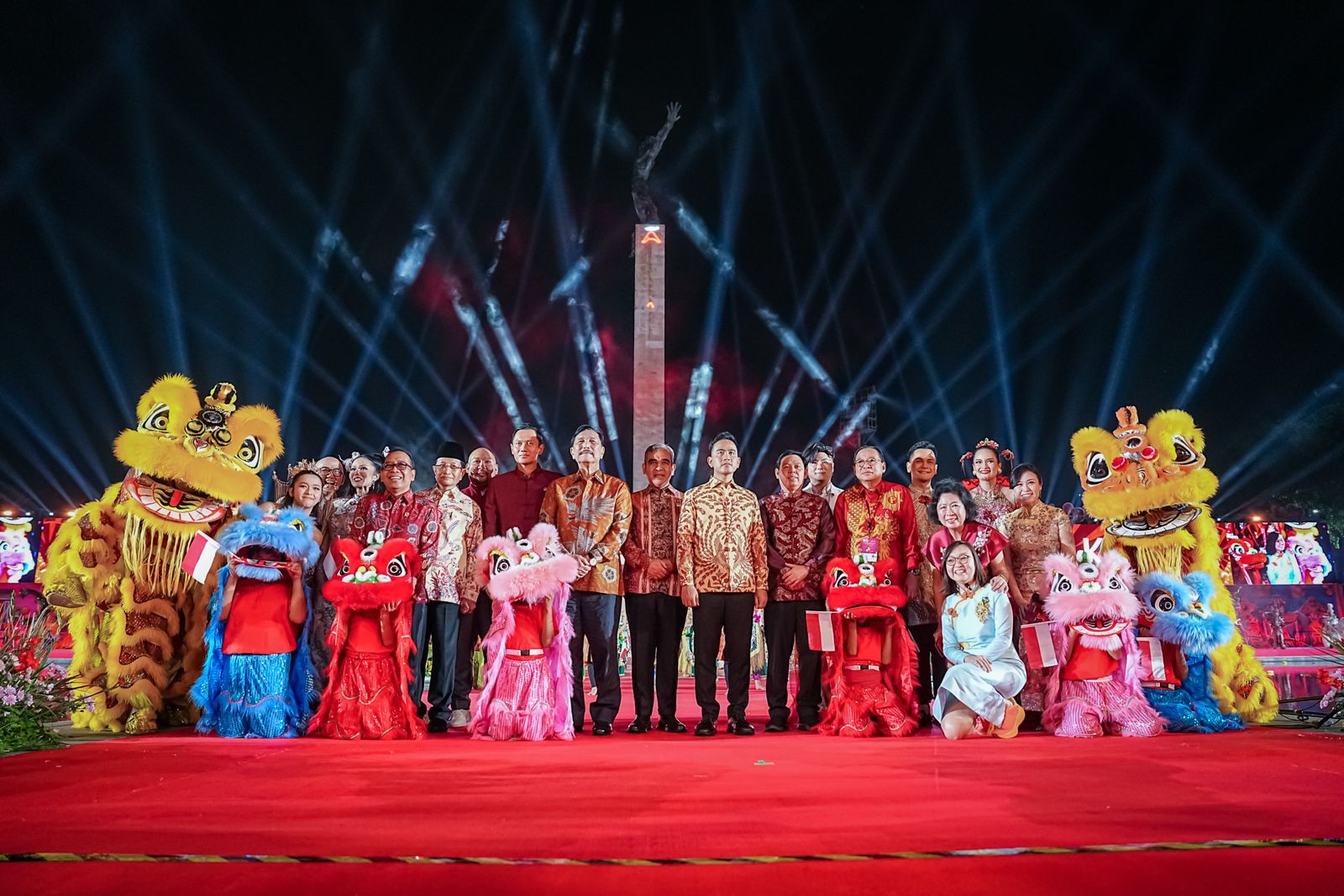 A group of people in traditional attire poses on a red carpet with lion dance costumes, under colorful lights at a festive event.