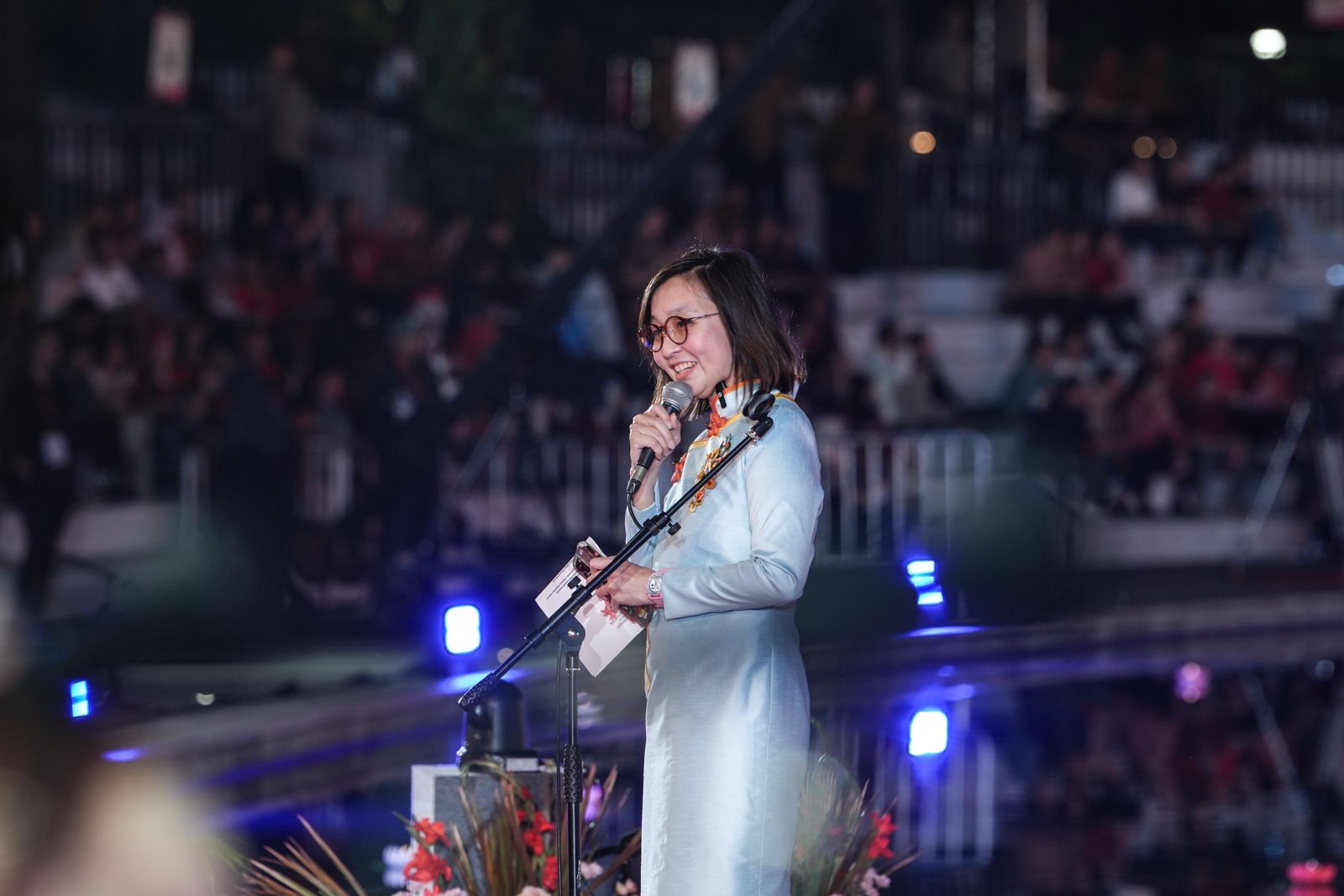 A woman in a light blue dress speaks into a microphone at an event, smiling at the audience in the background.