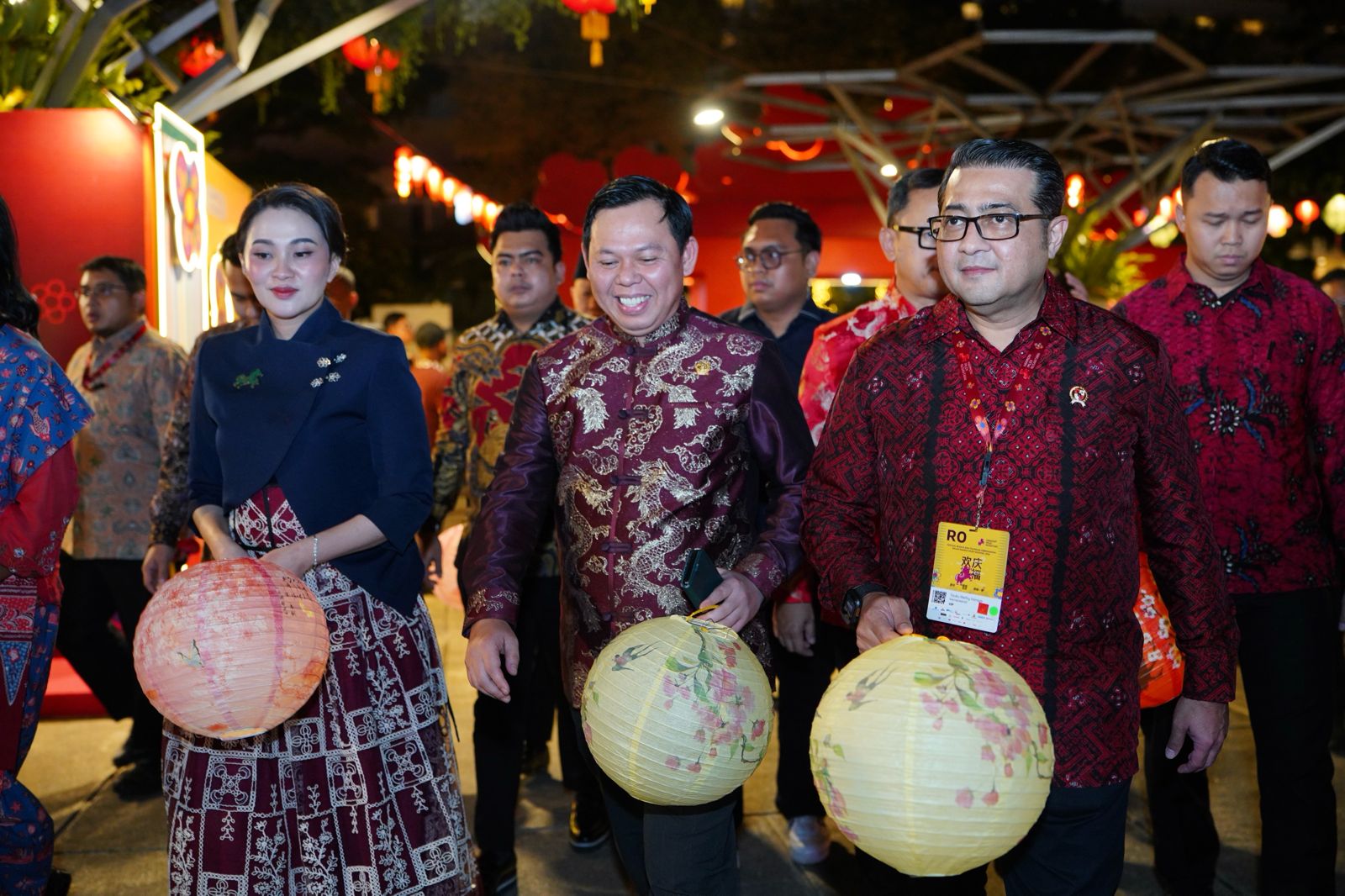 A group of people in traditional attire, holding lanterns, walking together at a festive night event. Colorful decorations are visible in the background.