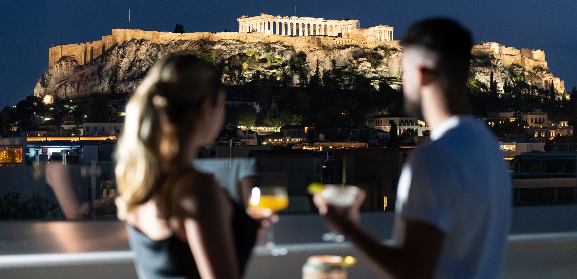 A couple drinking cocktails with Acropolis view by night