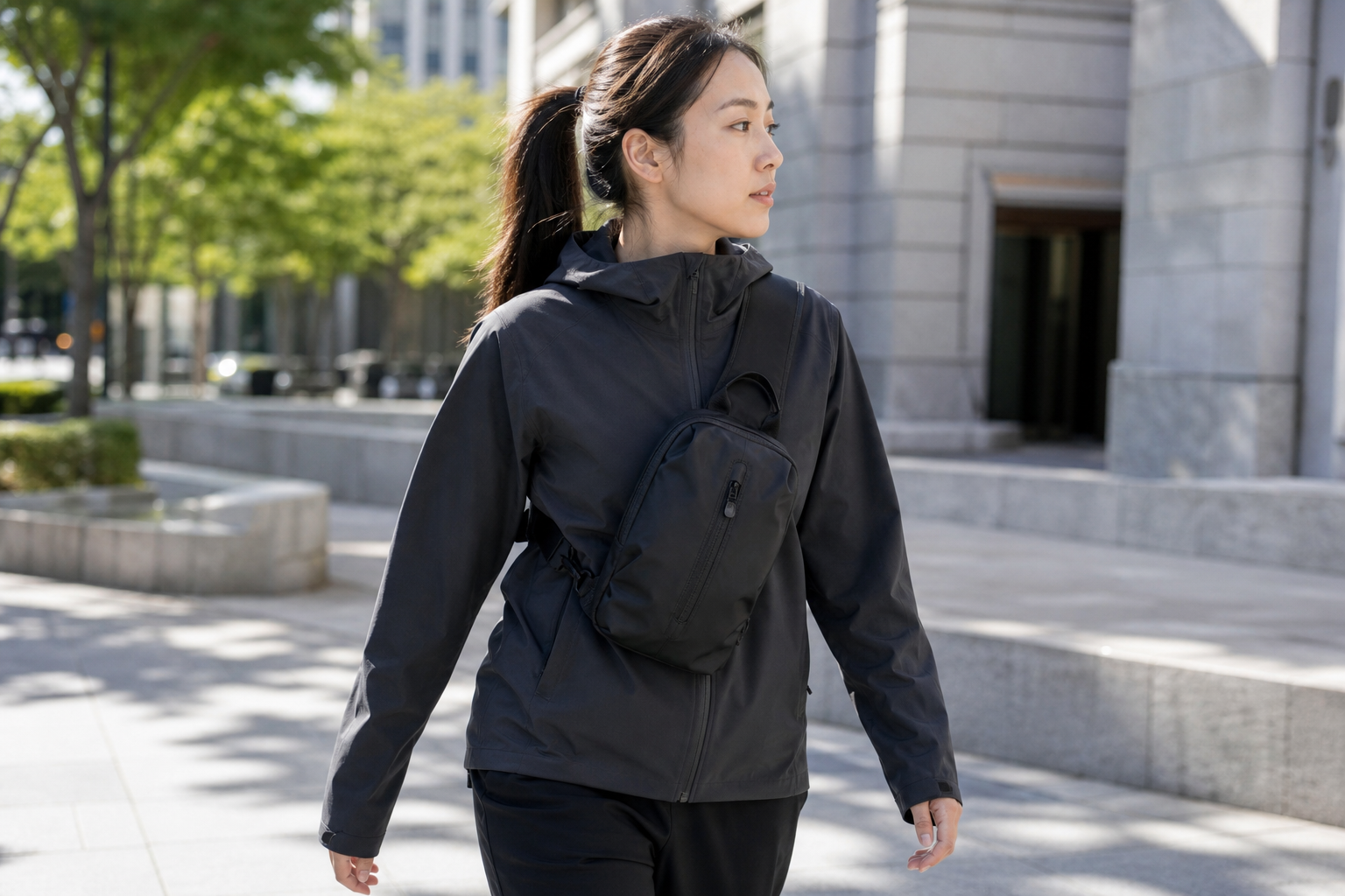 Woman walking through a sunlit urban plaza wearing a small black crossbody sling bag, hands free