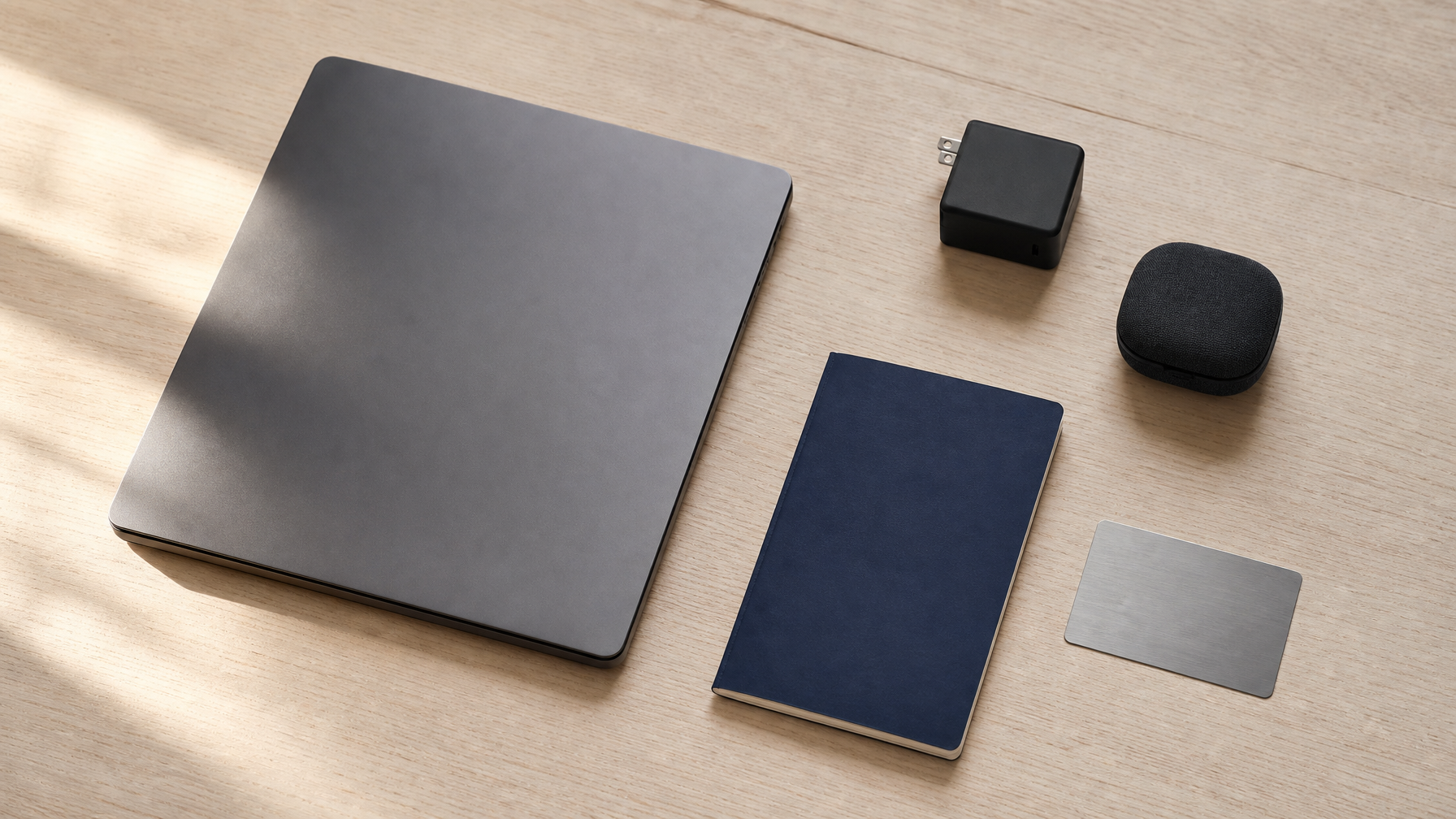 Overhead flatlay on pale wood: a closed laptop, compact GaN charger cube, dark fabric earbuds case, navy notebook, and brushed silver transit card arranged with intentional spacing