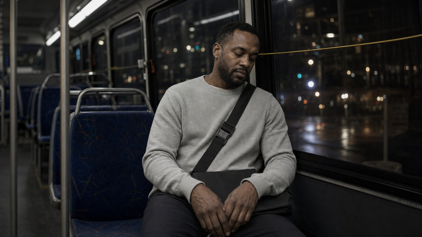 Man seated on a nearly empty night bus with a slim black laptop brief closed on his lap and city lights in the window