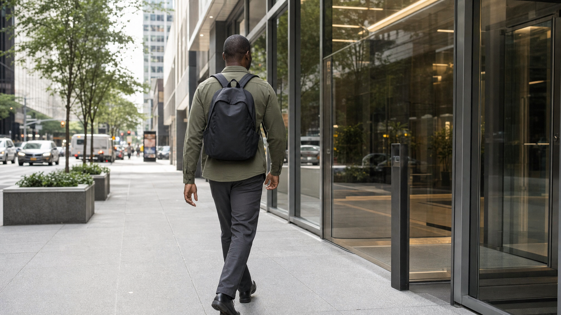 Man walking toward a glass office lobby with a closed black backpack on both shoulders