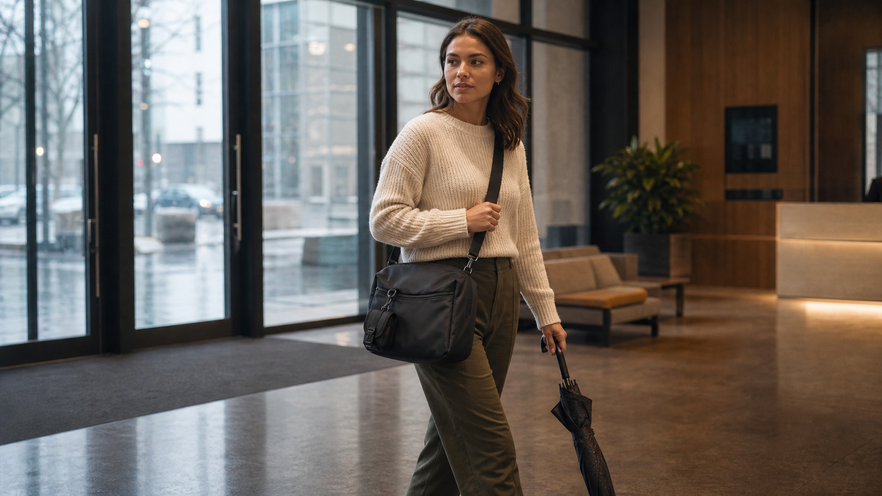 Commuter arriving in an office lobby on a damp morning with a closed black messenger bag and umbrella