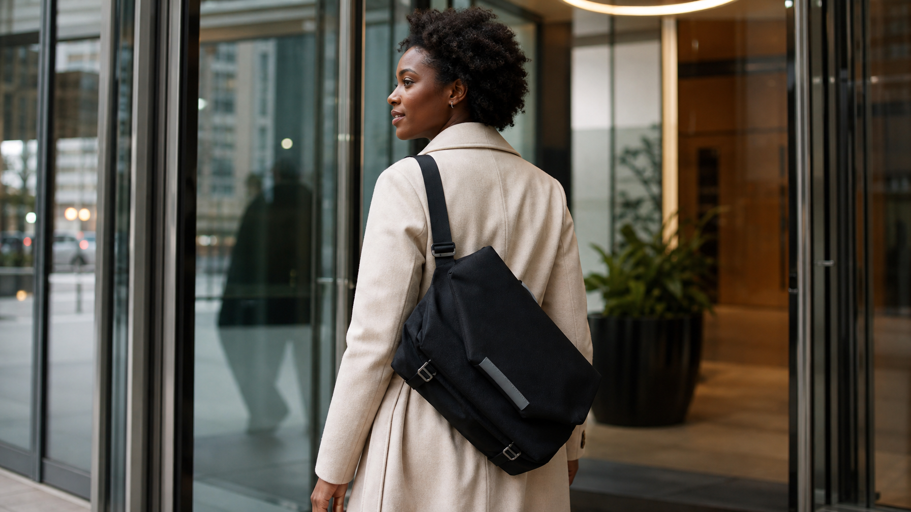 Black woman in her early 30s walking through an office building lobby with a medium black messenger on her shoulder