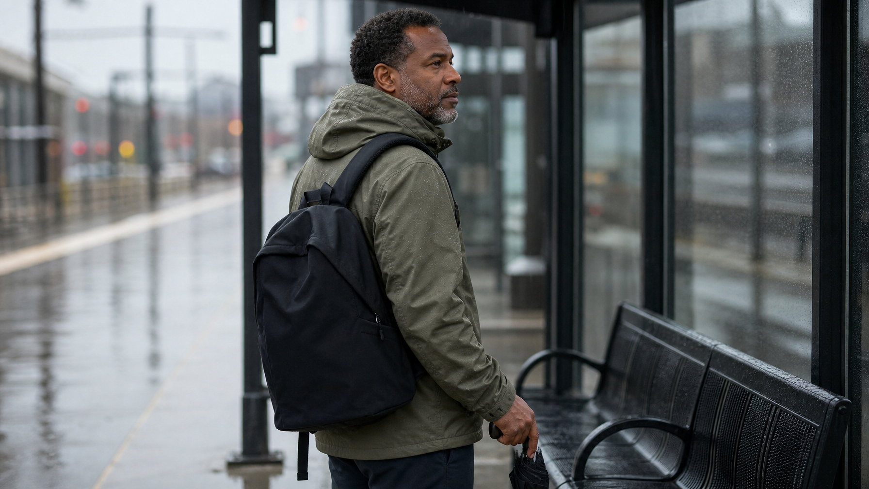 Commuter standing under a wet platform shelter wearing a slim black backpack with a compact umbrella in hand and an empty wet bench beside him