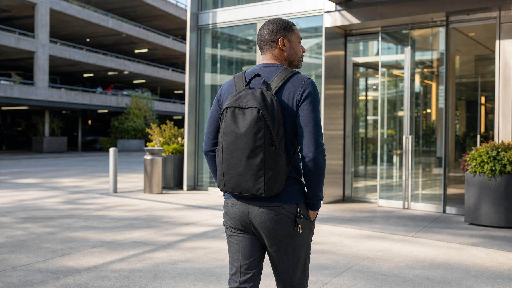 Man walking from a parking garage toward a glass office lobby with both backpack straps on and the pack closed