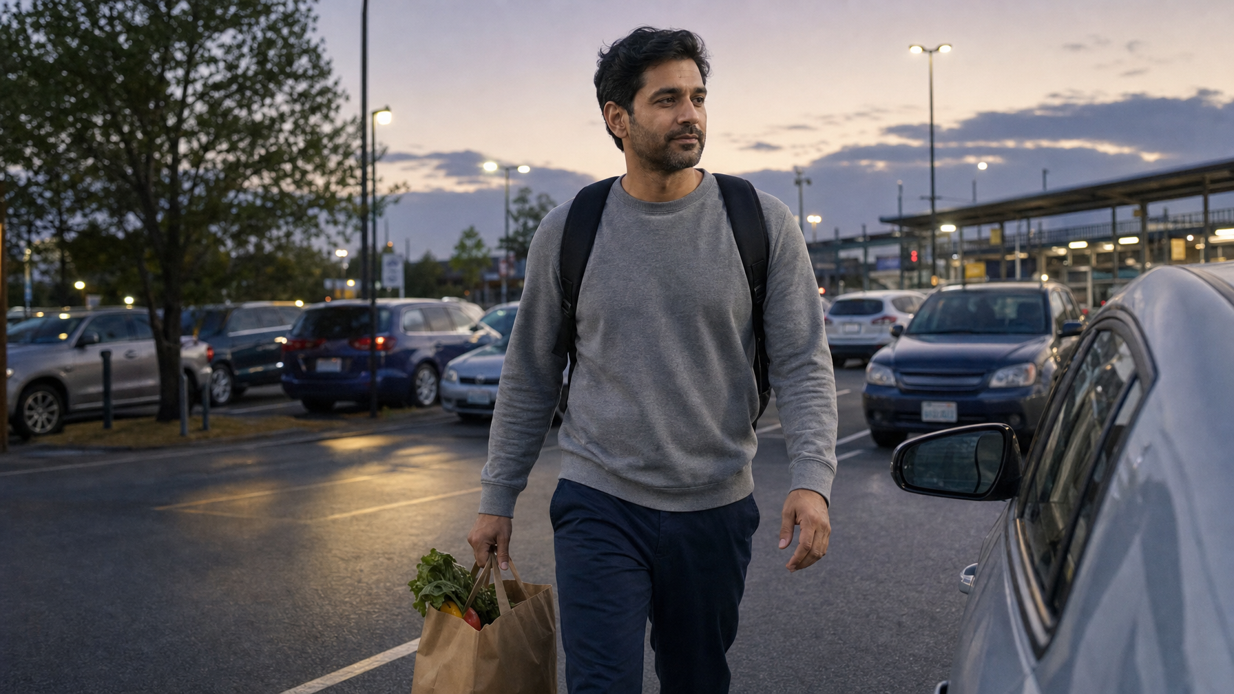 Man walking through a park-and-ride lot at dusk with both backpack straps on and a grocery bag in one hand